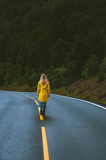 Escape it all woman walking on the road in forest traveling alone freedom concept vacations outdoor in Norway girl wearing yellow raincoat on the way