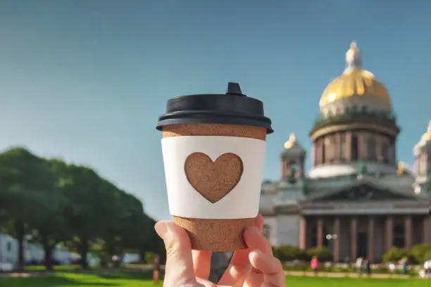 Photo of paper cup of natural coffee in woman's palm on background of St. Isaac's Cathedral in Saint-Petersburg on sunny day