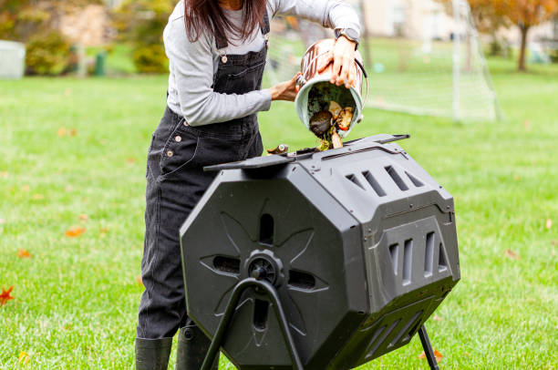 A woman is dumping a small bin of kitchen scraps into an outdoor tumbling composter in backyard garden stock photo