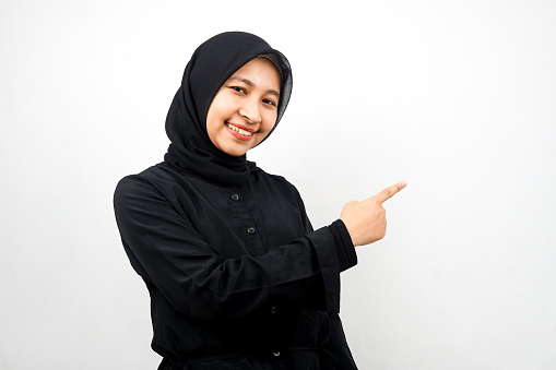 Beautiful young asian muslim woman with hands pointing empty space presenting something, smiling confident, enthusiastic, cheerful, looking at camera, isolated on white background, advertising concept