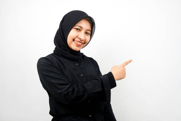 Beautiful young asian muslim woman with hands pointing empty space presenting something, smiling confident, enthusiastic, cheerful, looking at camera, isolated on white background, advertising concept stock photo