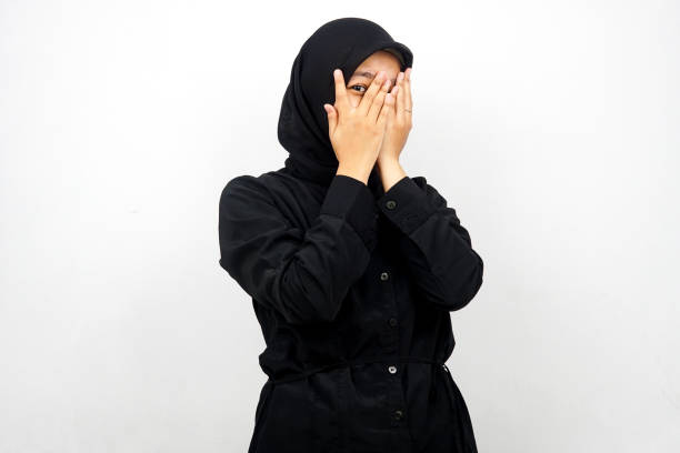 Beautiful young asian muslim woman with eyes peeking between fingers, isolated on white background stock photo