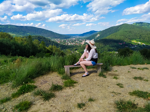 Happy girl in a straw hat, black and white blouse and blue shorts sitting on a bench with the mountain view. Calm and quiet wanderlust concept moment when person feels happiness and life.