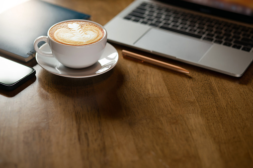 Focus on latte art coffee in white cup with computer laptop, mobile phone and blue note book on wooden table