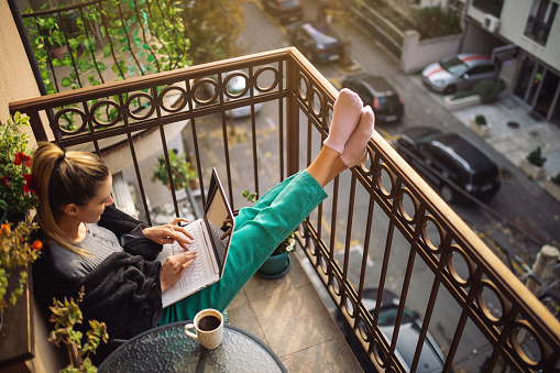 Beautiful young woman sitting on terrace, using laptop and drinking coffee at morning. Steam rising from coffee cup on the table. Cold morning, she is covering herself with warm blanket