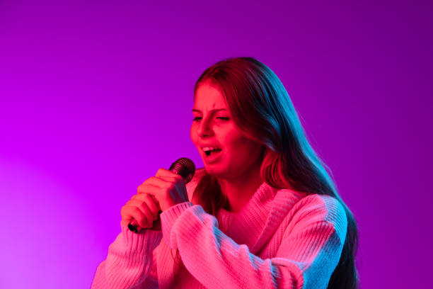 istock Young smiling beautiful girl singing with microphone isolated on purple studio background in neon pink filter. Concept of emotions, 1355617419