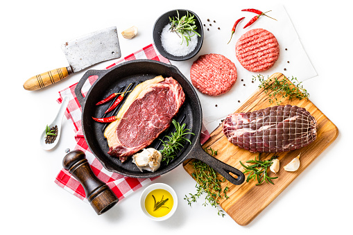 Top view of various kinds of raw meat such as a beef steak, a roast beef and burgers on a white background. The composition includes a cutting board, a kitchen axe,  a cast iron grill, salt, pepper and some aromatic herbs for seasoning raw meat.