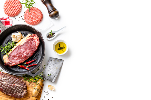 Top view of various kinds of raw meat such as a beef steak, a roast beef and burgers on a white background. All the objects are at the left of the image leaving a useful copy space at the right side. The composition includes a cutting board, a kitchen axe,  a cast iron grill, salt, pepper and some aromatic herbs for seasoning raw meat.