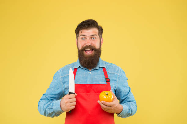 黄色いトマトの赤いエプロンのひげを生やした男。健康的で新鮮な食べ物を調理します。ハンサムな男性シェフが働いています。成熟した料理は野菜をカットします。キッチンで制服を着た� - minute maid beverages ストックフォトと画像