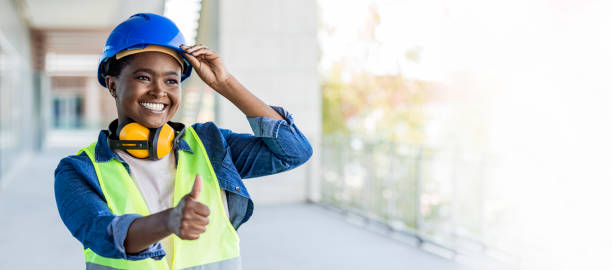 una mujer afroamericana de unos 30 años que trabaja en un sitio de construcción, usa un casco, gafas de seguridad y chaleco reflectante. - safety sign protective workwear factory fotografías e imágenes de stock