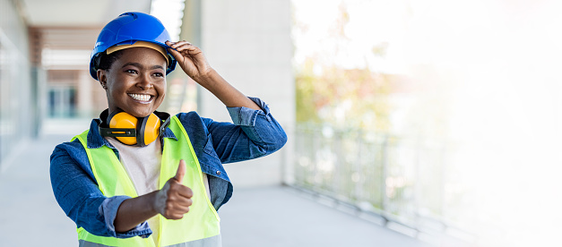 A African-American woman in her 30s working at a construction site, wearing a hardhat, safety goggles and reflective vest. She is looking over the camera with a confident expression, smiling, showing thumb up.