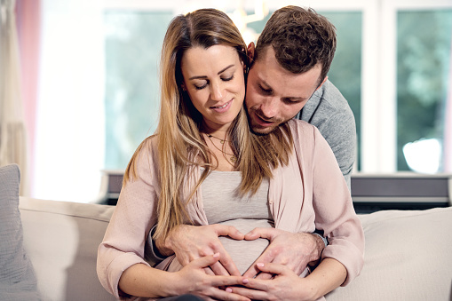 Happy young pregnant couple sits together on couch at home husband embracing wife gently holding her belly enjoying intimate moment