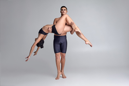 Two young graceful ballet dancers in black suits are posing over a gray studio background. Man in black shorts is holding a woman in a black swimwear. Ballet and contemporary choreography concept. Art photo.