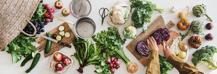 Fall vegetarian cooking background. Female hands cutting cabbage on wooden board over table with fresh seasonal vegetables, greens, fruit from grocery market, top view. Vegan, healthy, organic food