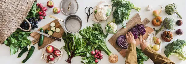 Fall vegetarian cooking background. Female hands cutting cabbage on wooden board over table with fresh seasonal vegetables, greens, fruit from grocery market, top view. Vegan, healthy, organic food