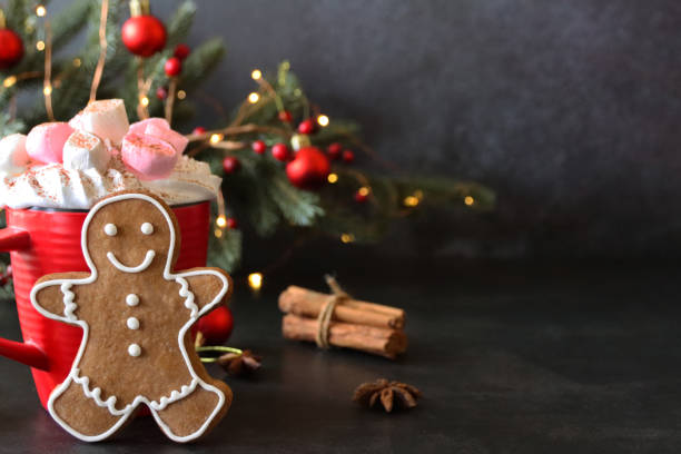 istock Image gingerbread man biscuit with white royal icing leaning on red hot chocolate mug topped by whipped cream, mini marshmallows, dried citrus orange fruit slices, cinnamon sticks, Christmas tree, illuminated fairy lights on black background, copy space 1355641995
