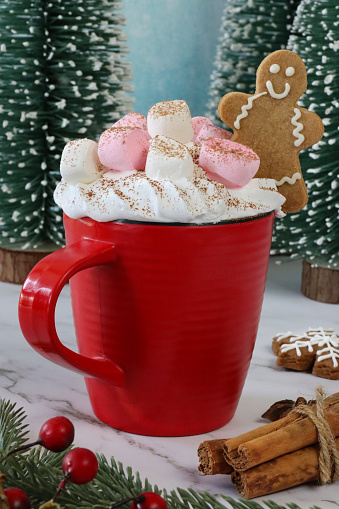 Stock photo showing close-up view of marble effect background with model fir trees surrounding red mug of hot chocolate topped, with whipped cream  marshmallows and a mini gingerbread man cookie.