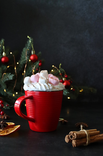 Stock photo showing close-up view of black background with a fallen mini Christmas tree covered in red and gold baubles, and illuminated fairy lights. Red mug of hot chocolate topped with whipped cream and mini marshmallows besides bundle of cinnamon sticks.