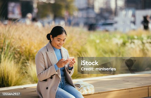 istock Smiling Asian Woman Sitting Outside and Looking at Her Phone 1355642927