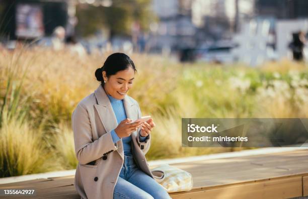 Smiling Asian Woman Sitting Outside And Looking At Her Phone Stock Photo - Download Image Now