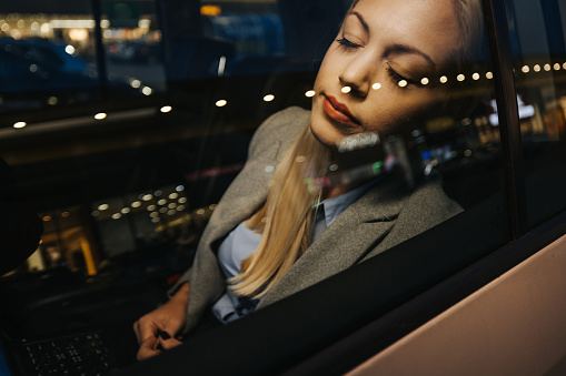 Young business woman sitting on back car seat and sleeping after working on laptop at night