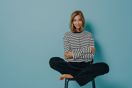 Relaxed young girl in striped shirt and jeans sitting with legs folded together in lotus pose on top of chair, huggs herself with hands pleasantly smiling, isolated on blue background with copy space