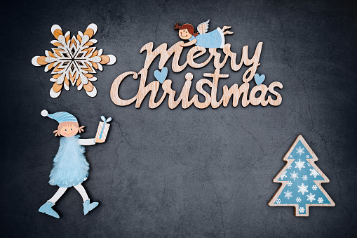 Flat lay Christmas card with dark background marbled effect with a wooden text of Merry Christmas with a smiling christmas doll holding a gift and a small blue wooden christmas tree and a wooden snowflake. Copy space in the centre. All articles are unbranded mass products from Chinese wholesalers. Color editing. Part of a series.