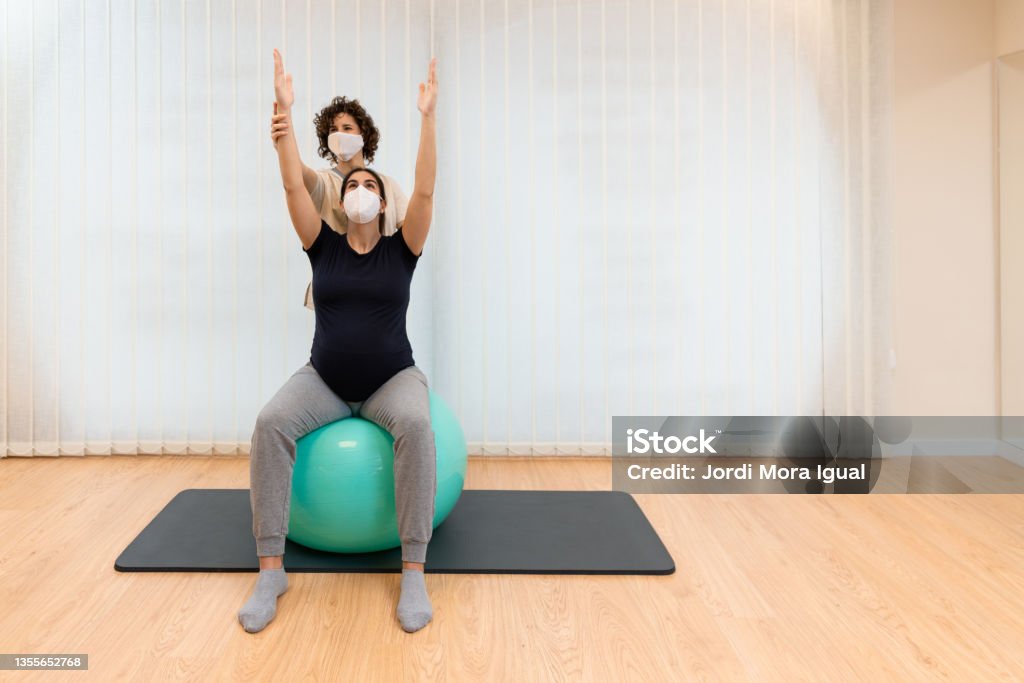 Physiotherapist helping to stretch the arms of a pregnant woman to exercise sitting on a fitness ball Front view of a physiotherapist helping to stretch the arms of a pregnant woman to exercise sitting on a fitness ball with copy space. Concept of pilates for pregnant women. Active Lifestyle Stock Photo