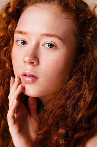 portrait of red-haired girl with long curly hair and cute freckles on beige background in the studio. Retouched photo. Skin care concept with pigmentation