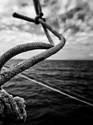 A black and white close-up detail shot of a knot on a rope. The shot is taken in the Mediterranian sea on a touristic yatch from a blue cruise. A dramatic sky with clouds and horizon over the water is visible in the background.