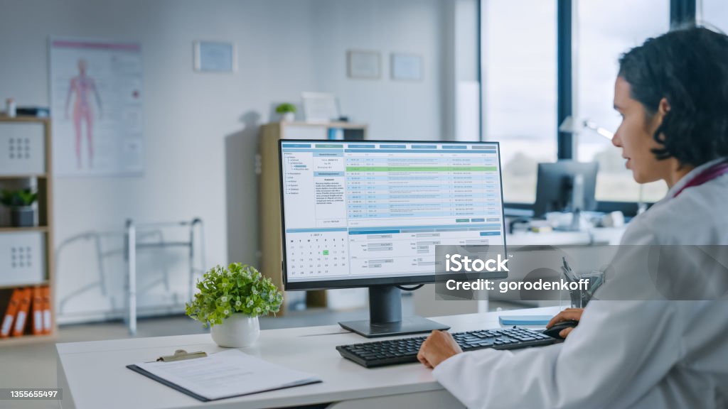 Experienced Female Medical Doctor Wearing White Coat Working on Personal Computer in a Health Clinic. Medical Health Care Professional Working with Test Results, Patient Treatment Planning. Computer Stock Photo