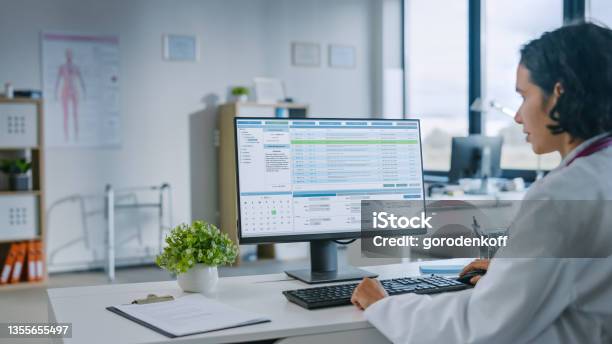 Experienced Female Medical Doctor Wearing White Coat Working On Personal Computer In A Health Clinic Medical Health Care Professional Working With Test Results Patient Treatment Planning Stock Photo - Download Image Now