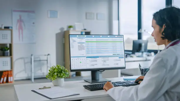 Photo of Experienced Female Medical Doctor Wearing White Coat Working on Personal Computer in a Health Clinic. Medical Health Care Professional Working with Test Results, Patient Treatment Planning.