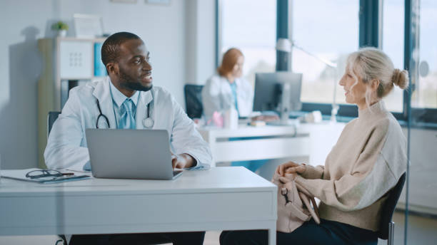 hausärztin liest die krankengeschichte einer älteren patientin und spricht mit ihr während der konsultation in einer gesundheitsklinik. arzt im laborkittel sitzt hinter einem laptop im krankenhausbüro. - healthcare and medicine smiling group of people lab coat stock-fotos und bilder