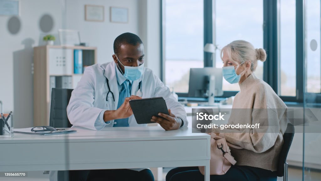African American Doctor in Protective Mask is Reading Medical History of Senior Female Patient During Consultation in a Health Clinic. Physician Using Tablet Computer in Hospital Office. Doctor Stock Photo