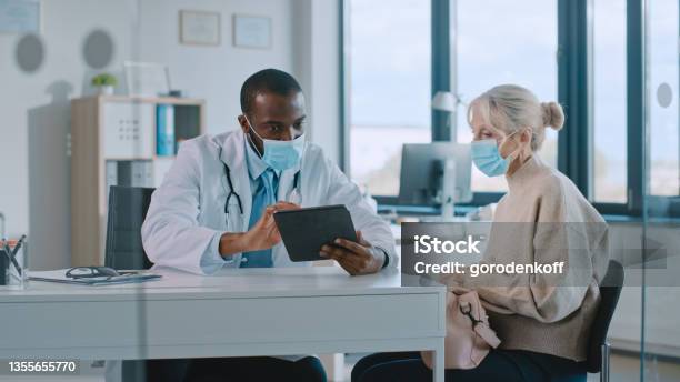 African American Doctor In Protective Mask Is Reading Medical History Of Senior Female Patient During Consultation In A Health Clinic Physician Using Tablet Computer In Hospital Office Stock Photo - Download Image Now