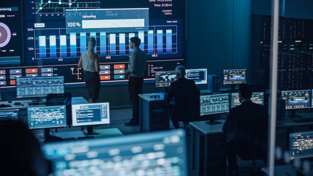 istock Two Software Engineers Talking Next to a Big Screen in a Modern Monitoring Office with Live Analysis Feed with Charts. Monitoring Room Big Data Scientists and Managers Sit in Front of Computers. 1355656495