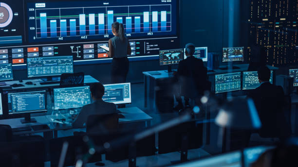 istock Software Engineers Working in a Room with a Big Screen in a Modern Monitoring Office with Live Analysis Feed with Charts. Monitoring Room Big Data Scientists and Managers Sit in Front of Computers. 1355656514