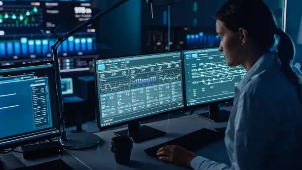 Female Software Engineer Working in a Modern Monitoring Office with Live Analysis Feed with Charts on a Big Digital Screen. Monitoring Room Big Data Scientists and Managers Sit in Front of Computers.