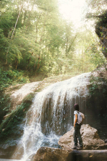 istock Woman tourist with a backpack walks next to the waterfall. 1355657280