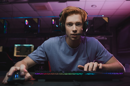 Waist up portrait of a handsome ginger man wearing headset sitting in front of the computer and being involved at the game