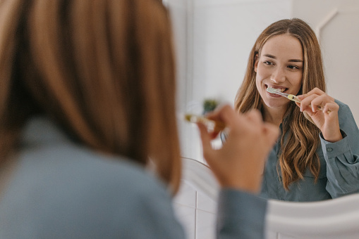 One woman, beautiful female looking in the mirror and brushing teeth in bathroom.