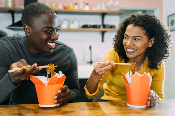 Smiling couple having lunch together at home. Shot of lovely african-american couple have good time together at home while eating Chinese food for lunch or dinner. Man and woman eating chinese takeout food in carton boxes at home. chinese takeout stock pictures, royalty-free photos & images