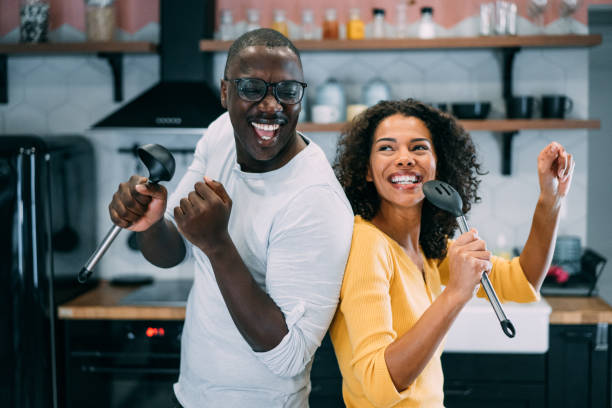 Fun in the kitchen. Shot of happy couple having fun during cooking in the kitchen at home. Lovely african-american couple holding kitchen utensil and singing during a cooking. african american couple stock pictures, royalty-free photos & images
