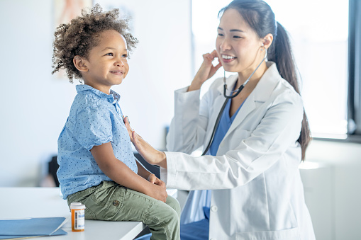 A female doctor of Asian decent holds her stethoscope to a little boys chest as she listens to his heart.  The little boy is dressed casually and smiling as he sits still for the appointment.
