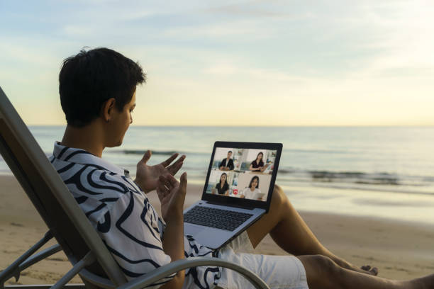 uomo d'affari asiatico che ha videoconferenza a distanza con il suo team di lavoro in spiaggia durante le vacanze in vacanza. - office water business meeting foto e immagini stock