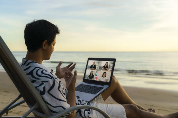 asian business man having remote video conference call with his business team at the beach during vacation in holiday. - on beach laptop working imagens e fotografias de stock