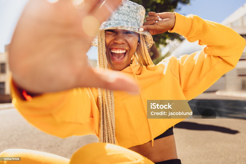Young woman blocking the camera Young woman blocking the camera with her hand. Happy young woman smiling at the camera cheerfully while sitting outdoors on a summer's day. Vibrant young woman wearing stylish long braids. Candid Stock Photo