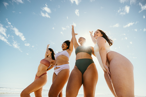 Celebrating spring break. Group of happy female friends cheering with their arms raised while wearing swimwear at the beach. Carefree young women having fun and enjoying their vacation.