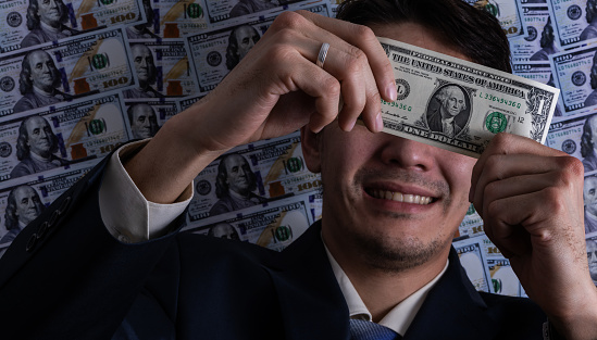 Smiling businessman almost covering his face and holding a 1 American dollar banknote against the background of 100 American dollar bills